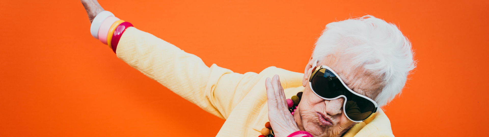 An elderly woman with white hair dabbing, wearing sunglasses and a yellow outfit, with an orange background.