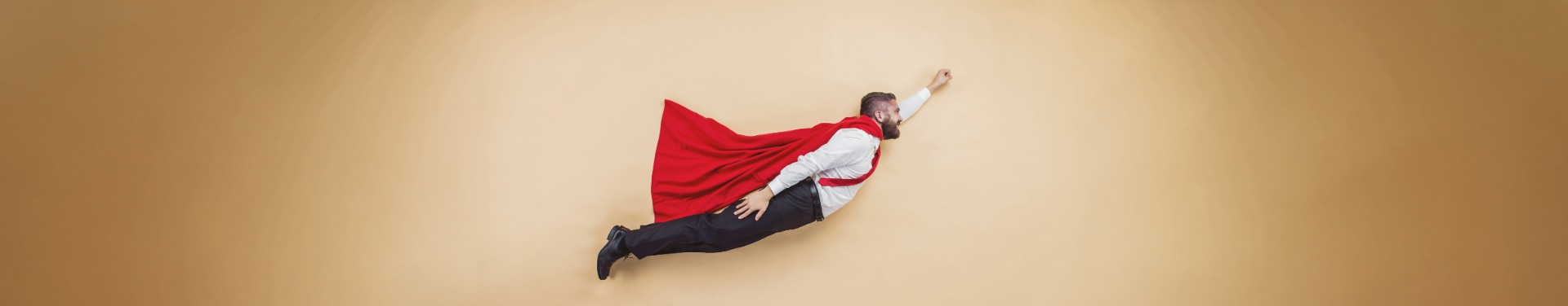 A person dressed in formal attire with a red cape poses as if flying against a light beige background, with his arm outstretched forward in a superhero pose.