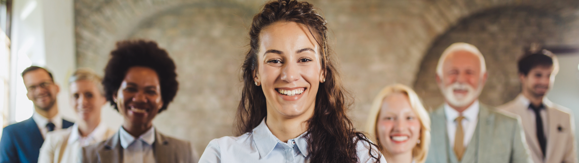 A smiling woman stands at the front of a group of diverse colleagues, all dressed in business attire.