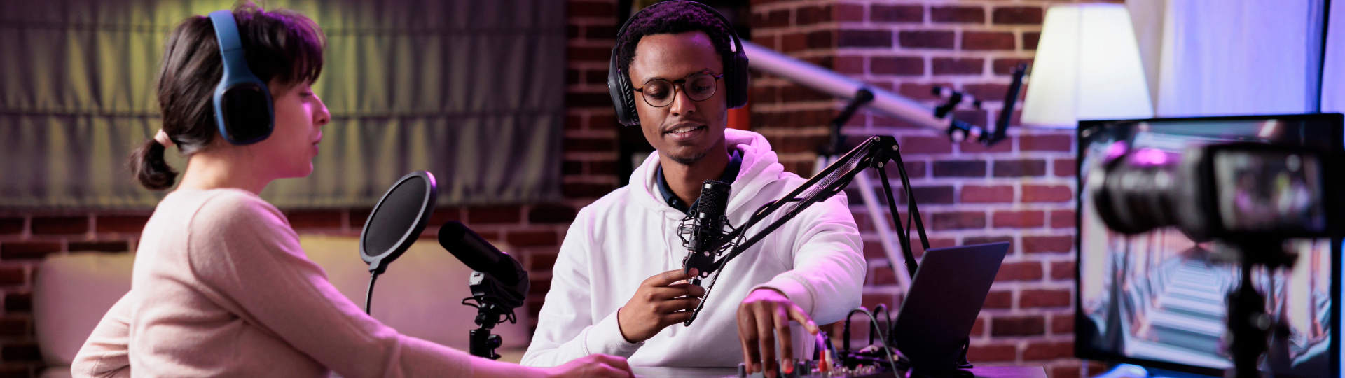 Two podcast hosts in a studio, one adjusting audio equipment, the other speaking into a mic.