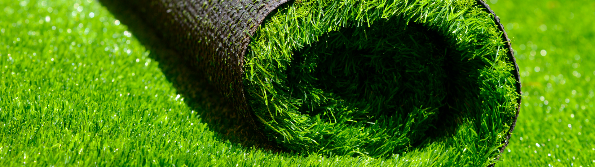 A close-up image of a roll of artificial (synthetic) grass, partially unrolled on a vibrant green synthetic lawn.