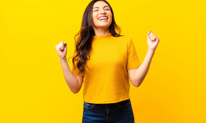 A smiling woman in a yellow shirt excitedly pumping her fists against a bright yellow background.