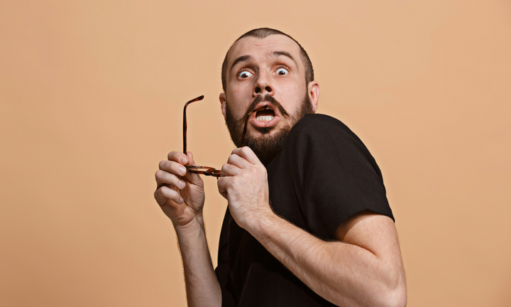 A bearded man wearing a black shirt holds his glasses, looking surprised and leaning back in shock.