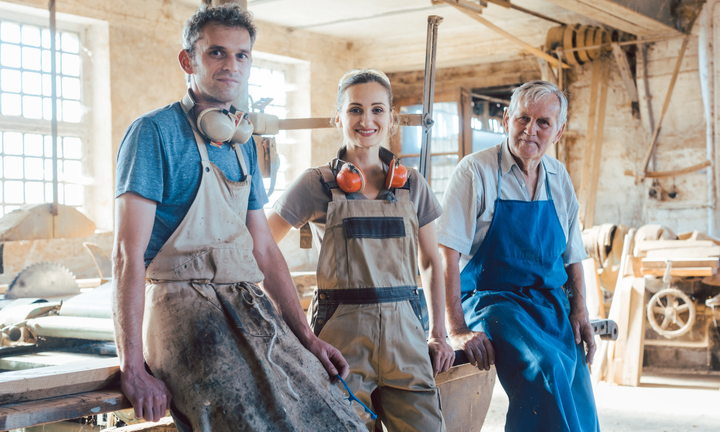 A group of three craftspeople in a workshop: two men and a woman wearing aprons, smiling confidently.